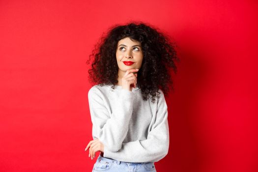 Pensive stylish woman with curly hair, red lips, looking thoughtful at upper left corner logo and smiling, standing against studio background.