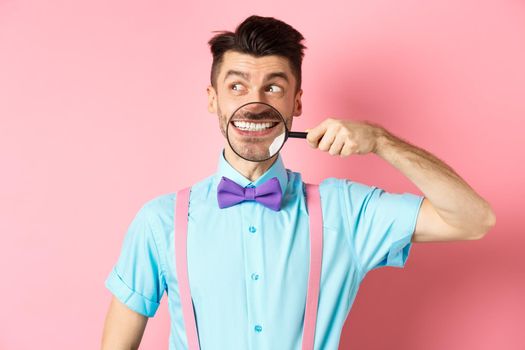 Funny man in bow-tie smiling, showing teeth with magnifying glass and looking aside at logo, standing over pink background.