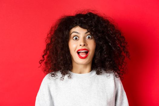 Close-up portrait of excited woman with curly hair, scream surprised and amazed, checking out special deal, standing on red background