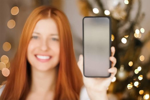 Christmas phone call and holiday greeting concept. Happy smiling woman showing screen of mobile smartphone, xmas decoration on background