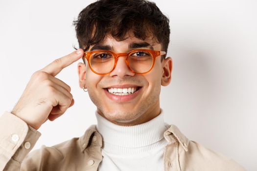 Headshot of smiling happy man showing new eyewear frame, pointing at glasses and looking satisfied, standing on white background