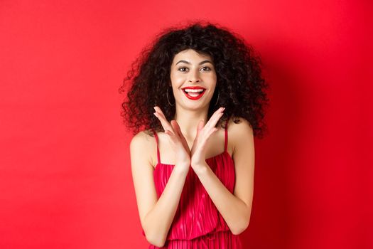 Cheerful young woman with curly hair, wearing evening dress, clap hands and smiling, praise good performance with applause, standing against red background