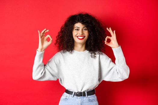 Confident pretty girl with curly hairstyle, showing okay gestures and smiling, approve and agree with you, praising excellent choice, standing satisfied on red background