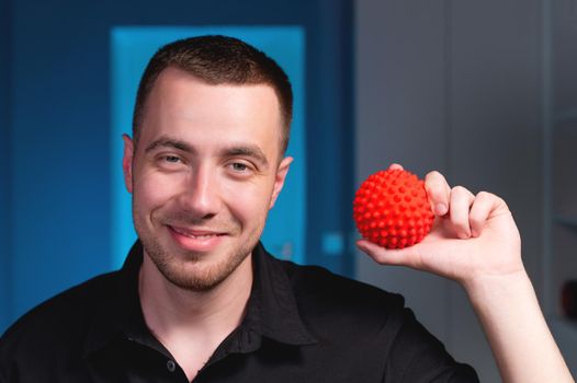 Portrait of a young Caucasian physiotherapist masseur on a blue background with a red massage ball in his hands. Smiling and looking at the camera. Self-massage and self-treatment concept