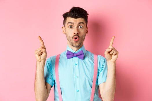 Impressed young guy with bow-tie point fingers up, showing top promo offer and say wow, standing over pink background