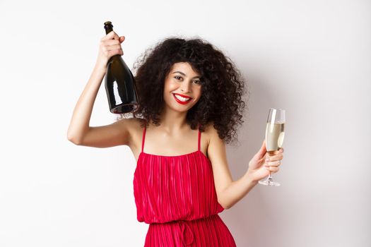 Happy woman partying on valentines day holiday, dancing with glass and bottle of champagne, wearing red dress, smiling on white background
