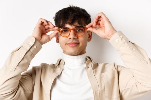 Close-up of stylish hipster guy trying eyewear at optician store, put on glasses and smiling, standing on white background
