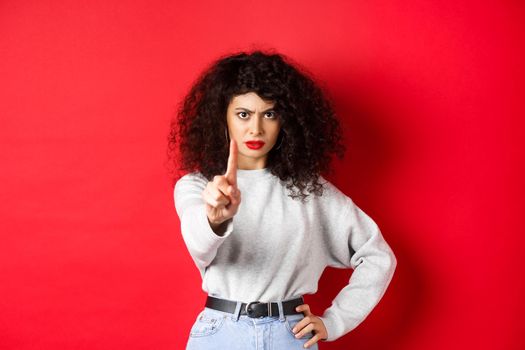 Serious confident woman say no, extend one finger to stop you, prohibit something bad, standing determined against red background