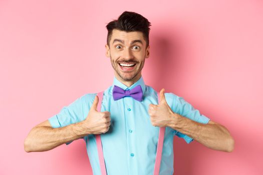 Happy young man showing thumbs up and smiling, saying yes, agree with something cool, recommending excellent deal, standing upbeat on pink background