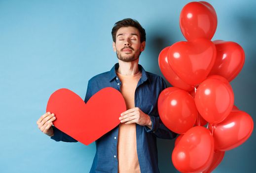 Valentines day concept. Man dreaming of true love, holding red heart cutout and standing near romantic balloons, blue background