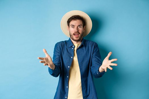Surprised young man in summer hat, raising hands up staring with disbelief and amazement at camera, impressed with big event, standing on blue background