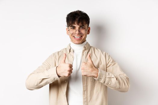 Cheerful hipster guy in glasses showing thumbs up and smiling, like good product, recommend or praise advertisement, standing on white background