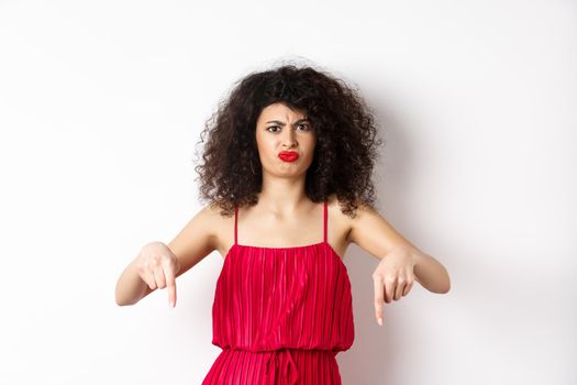 Disappointed caucasian woman in red dress complaining on spoiled date, frowning and grimacing, standing over white background