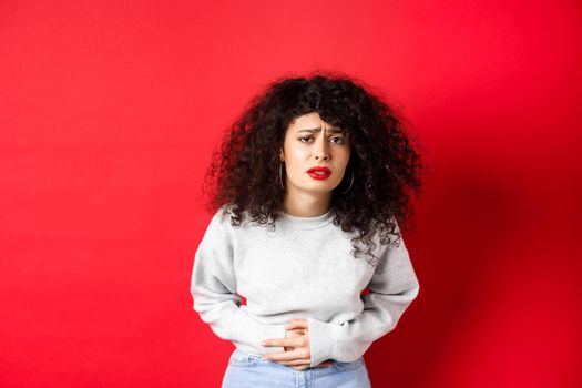 Image of young woman having stomach ache, bending from pain and complaining on painful menstrual cramps, standing on red background