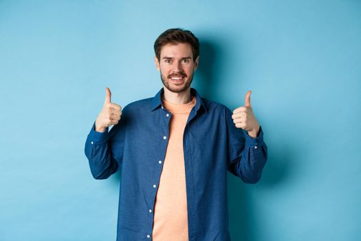 Handsome smiling man being supportive, showing thumbs up in approval, praise good work, standing happy on blue background