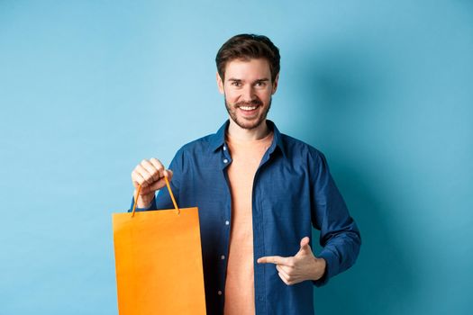 Smiling caucasian man pointing finger at orange shopping bag with bought items, standing on blue background
