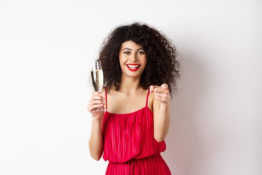 Beautiful curly woman in red dress, partying or having romantic date, holding glass of champagne and pointing at you, inviting person, standing on white background