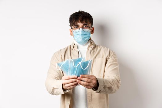 Health, covid and quarantine concept. Image of young guy handing out medical masks for preventive measures, helping during pandemic, standing on white background