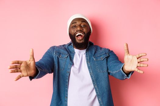 Happy Black man stretching hands, reaching for hug or take something, smiling pleased, standing in beanie and denim shirt over pink background