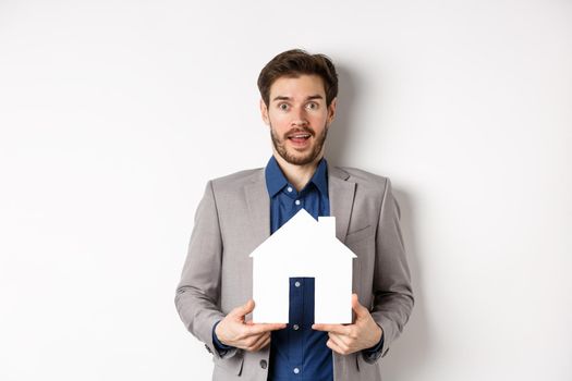 Real estate. Excited business man in suit holding paper house cutout and looking amazed at camera, buying apartment, white background