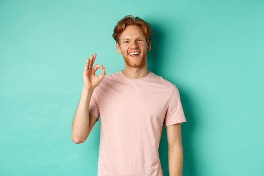 Handsome young bearded man in t-shirt showing Ok sign, smiling with white teeth and saying yes, agree with you, standing over turquoise background