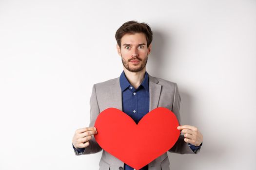 Excited caucasian man in suit looking at camera, holding big red heart cutout on valentines day, standing over white background