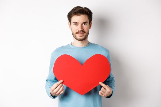 Smiling young man holding valentines heart cutout and looking at camera, waiting for true love girlfriend, standing over white background