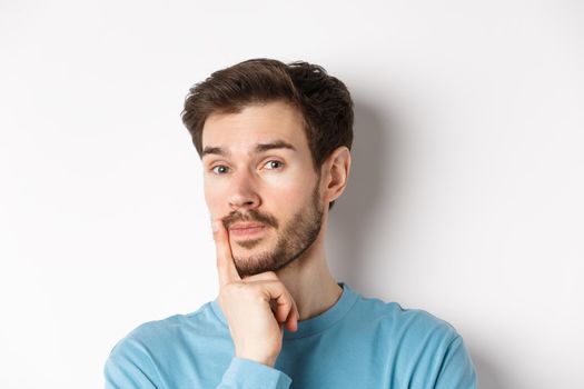 Image of thoughtful young man with beard, making choice, touching lip and looking pensive at camera, deciding something, white background