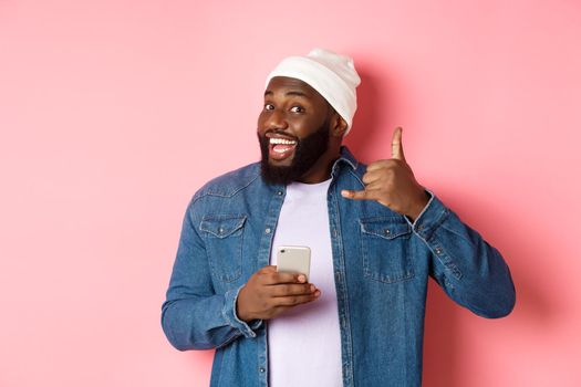 Happy Black man showing call me sign, making phone gesture and smiling, holding smartphone, standing in beanie and denim shirt over pink background