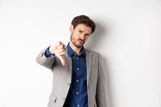 Negative emotions. Disappointed businessman frowning, showing thumb down to disapprove product, dislike something bad, stading on white background