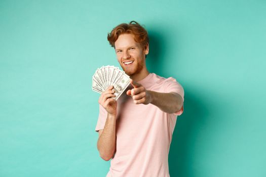Handsome and confident redhead guy pointing finger at you, showing money dollars and smiling, concept of earning cash and investment