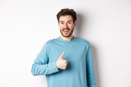 Cheerful young man agree and say yes, show thumb up in approval, smiling happy at camera, standing over white background