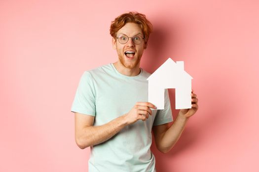 Real estate. Cheerful redhead man buying property, showing paper house cutout and smiling amazed, standing over pink background