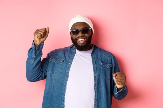 Happy Black man in beanie an sunglasses rejoicing, dancing with happy face, standing over pink background