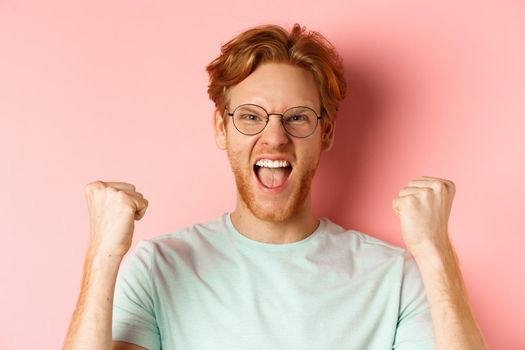 Close up of satisfied lucky redhead man winning, shouting from joy and making fist pump, celebratig victory, standing like champion over pink background