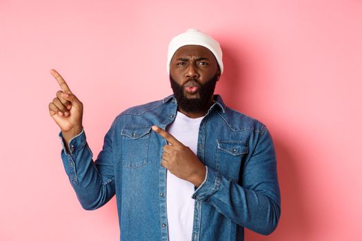 Confused and displeased african american man grimacing, cringe from disappointment and pointing fingers at upper left corner, bad promo, pink background