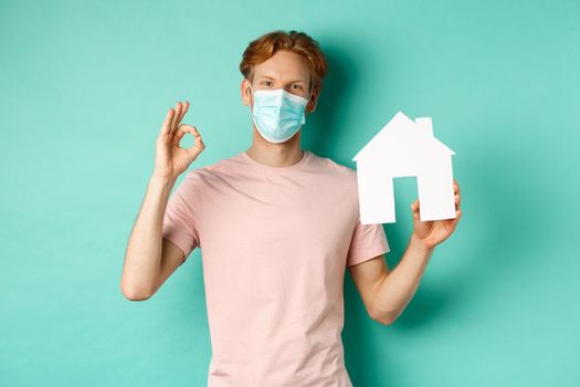 Covid-19 and real estate concept. Cheerful guy in face mask showing house cutout and okay sign, standing over turquoise background