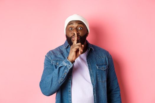Handsome young black guy in beanie and denim shirt prepare surprise, hushing softly and looking at camera, asking keep quiet, pink background