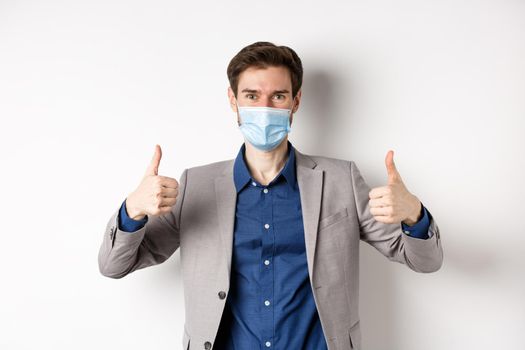 Covid-19, pandemic and business concept. Cheerful man in suit and medical mask using preventive measures in office, showing thumbs up, white background