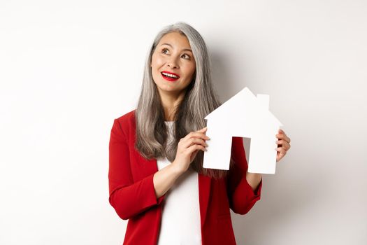 Dreamy senior woman thinking of buying property, showing paper house cutout and looking at upper left corner, standing over white background