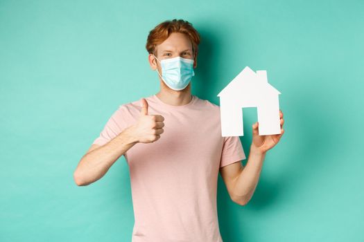 Covid-19 and real estate concept. Cheerful guy in face mask showing house cutout and thumbs-up, standing over turquoise background