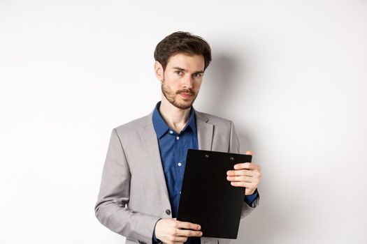 Young male ceo manager holding clipboard with documents and looking at camera, inspecting office, standing on white background