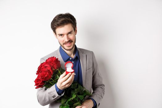 Romantic man with boquet of red roses asking to marry him, holding engagement ring and looking confident at camera, standing in suit over white background