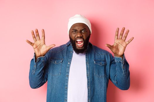 Angry african-american guy in beanie, scare you, roaring and screaming, showing hands, standing ove rpink background