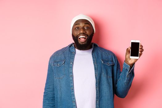 Handsome hipster guy in beanie and denim shirt smiling, showing mobile phone screen with happy face, recommend app, standing over pink background