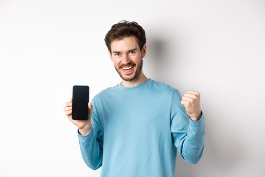 Image of happy man showing empty smartphone screen and celebrating, smiling with rejoice and fist pump, achieve online goal, standing over white background