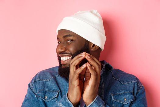 Close-up of devious african-american male model having an idea, scheming something, steeple fingers and smiling sly, standing over pink background