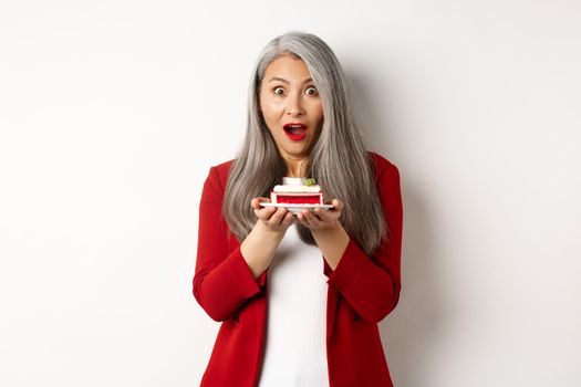 Surprised asian middle-aged woman holding piece of tasty cake, looking amazed at camera, standing over white background