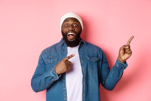 Excited and amazed Black man showing awesome offer, pointing fingers right at copy space, standing in hipster beanie and denim shirt on pink background