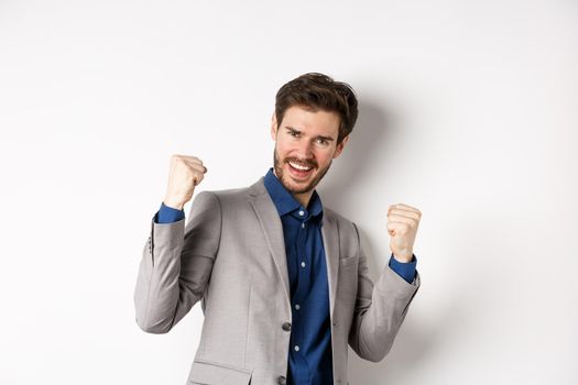 Lucky businessman winning money prize, say yes and smiling excited, make fist pump sign to celebrate victory, triumphing in suit on white background